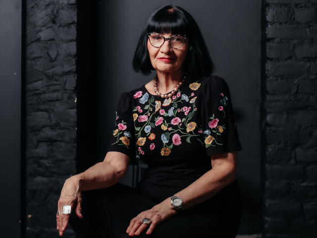 TiniaMartin, an older woman with black bob, glasses, and floral black top, sitting with legs crossed, wearing jewelry, black pants, and a watch, in a dark setting.