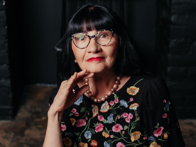 TiniaMartin has black hair, glasses, red lipstick, and a pearl necklace. She's wearing a floral dress, touching her chin with a hand. Her ass is not visible.