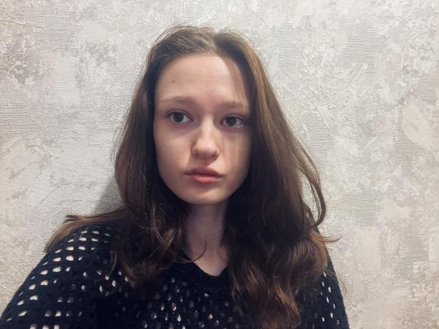 CharissaLeister has a sad, neutral expression with long brown hair, wearing a black, see-through knit top, showing her small, bare tits against a textured, gray wall.