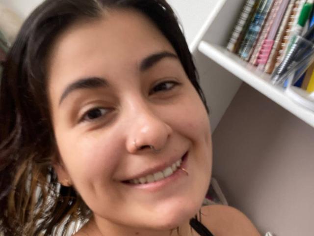 NyxVale's smiling face with brown eyes, dark hair, nose ring, and lip piercing. She's wearing a black top, and there's a bookshelf in the blurry background.