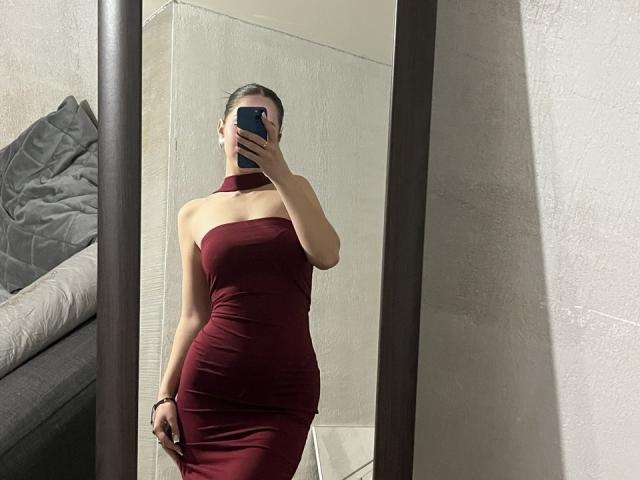 JudiBeedoo in a tight, strapless red dress, holding a phone, covering her face, with her ass and tits slightly visible, standing in front of a mirror.