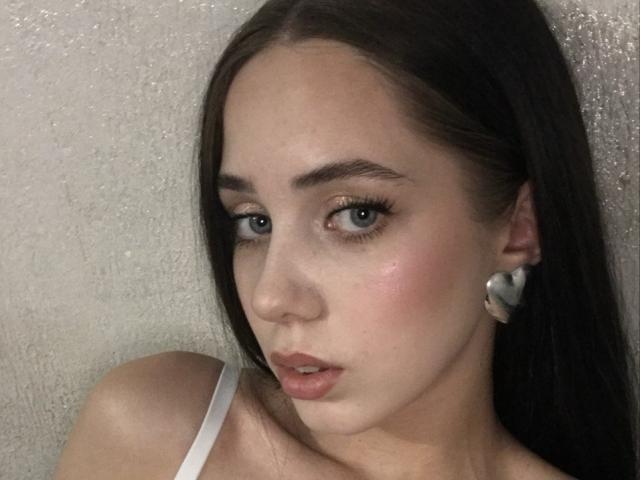 JudiBeedoo has pale skin, brown eyes, and long brown hair. She's wearing a white strap and small white earrings, with a slight blush on her cheeks and parted lips.