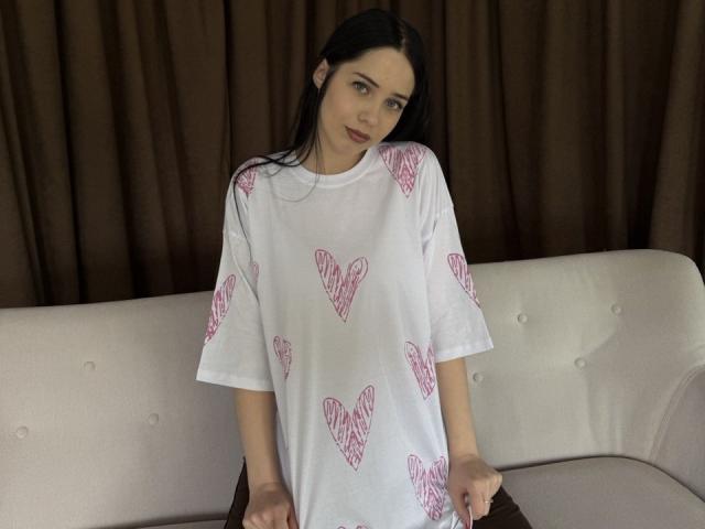 JudiBeedoo with long black hair, wearing a white shirt with pink heart patterns, standing in front of a beige couch, looking slightly to the side, smiling softly.