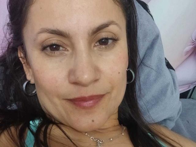 SolaraDubois has brown eyes, dark brown hair, a nose ring, hoop earrings, and a small mole on her right cheek. She's smiling with pink lipstick and a silver necklace.