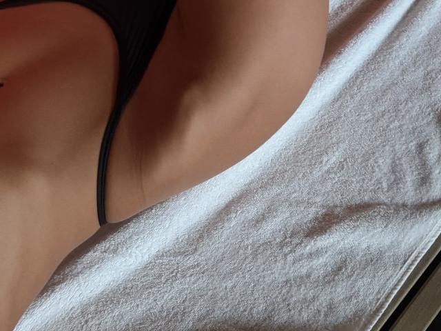 Fridaa's bare, tanned, smooth, curvy lower belly and black thong are visible on a white, textured towel, with sunlight casting soft shadows on her skin.