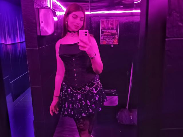 DolLemon com luzes roxas, vestindo um corpete preto, um colar e uma saia preta com padrões brancos, tirando uma selfie no espelho, mostrando os seios e um sorriso adorável..