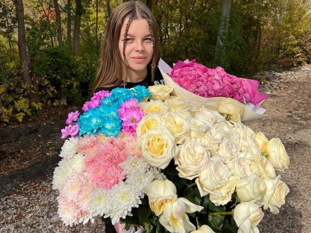 RhonaBells che tiene un bouquet di fiori colorati, tra cui rose rosa, blu, bianche e gialle, e garofani rosa, in piedi su un sentiero di ghiaia in una foresta..