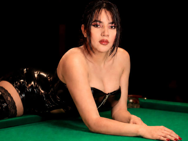 AliceCarusso, with black latex dress, lying on green pool table, her tits visible, black lace thigh-highs, red lipstick, and dark hair, looking at camera, hands on table.