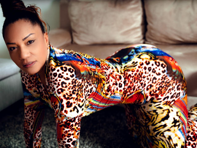 KrytalOwen, con un sedere prosperoso e sensuale, indossa un abito colorato con stampe di leopardo e motivi tribali, è inginocchiata su un tappeto e guarda la fotocamera con un sorriso leggero e sicuro..