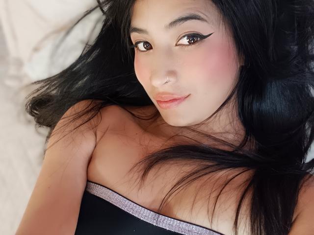 NaiaSanders with long black hair, brown eyes, and pink blush, lying down, wearing a black strap top, nose pierced, subtle smile, looking at camera. Boobs partially visible.