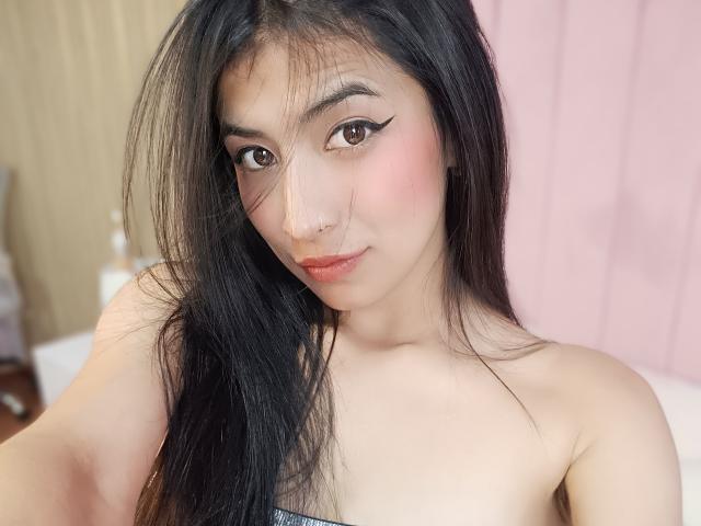 NaiaSanders with long black hair, brown eyes, pink blush, and winged eyeliner, smiling softly, showing her nose ring and bare shoulders, with her small tits visible.