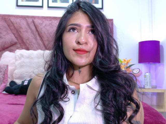 LuciadelaVega with long black wavy hair, red lipstick, nose piercing, white sleeveless shirt, small tattoo under chin, purple lamp, pink bed, white pillow, and framed pictures in background.