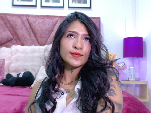 LuciadelaVega with long black wavy hair, red lipstick, tattoos on her neck and arm, gold necklace, white sleeveless top, sitting on a bed with pink and white pillows.