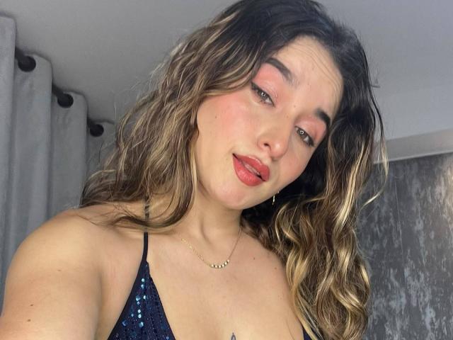 MilaGonz with wavy brown hair, blue sequined top, pink lipstick, slightly parted red lips, gold necklace, and a subtle smile. Her tits are small and her pussy is not visible.
