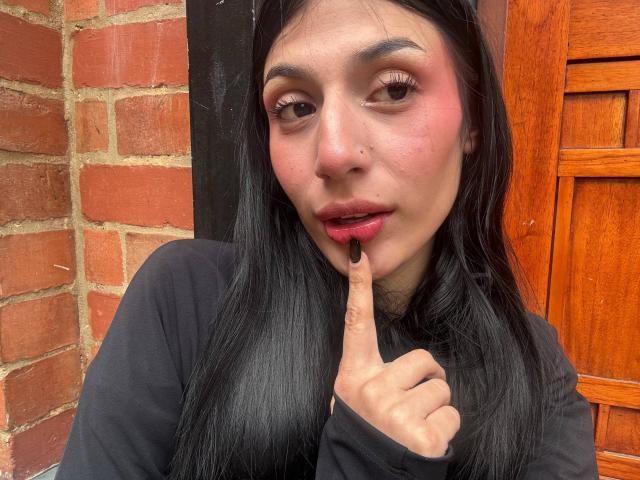SophieBeaumont with long black hair, pink eyeshadow, and black lipstick, touching her pink-tinted lips with a finger, wearing a black shirt, standing in front of a brick wall.