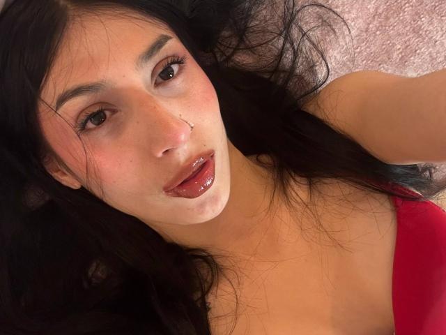 Sophie Beaumont, met lang zwart haar, bruine ogen, een neusring, glanzende rode lippen, lichte sproeten, draagt een rode top en ligt plat, met een zachte, roze achtergrond. Borsten zijn gedeeltelijk zichtbaar..