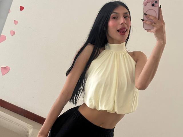 SophieBeaumont with long black hair, tongue out, wearing a white halter top and black pants, holding a pink phone, showing her small tits and flat stomach.
