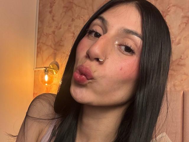 SophieBeaumont with long black hair, nose piercings, and pouty lips, making a kissing face, wearing a pinkish top, glowing light in background.