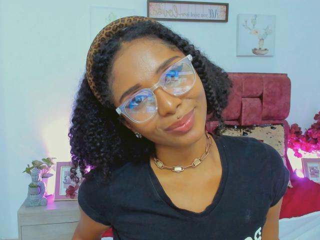 MeganWlliams with curly black hair, leopard print headband, clear glasses, black shirt, gold necklace, nose ring, and pink lips, smiling slightly. She's looking at the camera.