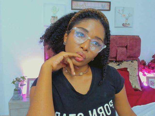 MeganWilliams with curly black hair, wearing clear glasses, leopard print headband, black shirt, and gold necklace, pouting with finger on her chin, looking serious.