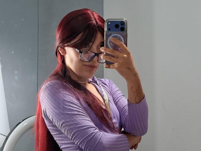 AbbyAndMichelross with red hair, glasses, and a purple ribbed top, holding a phone, showing tattoos on her arm, taking a mirror selfie, looking cute and confident.