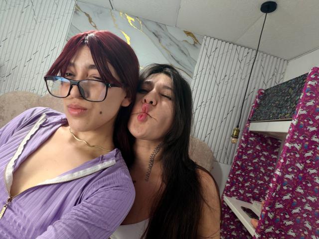 AbbyAndMichelross with red hair, glasses, and purple zip-up, and AbbyAndMichelross with black hair, kissing AbbyAndMichelross's cheek, showing tattoo, in a room with pink patterned shelf.