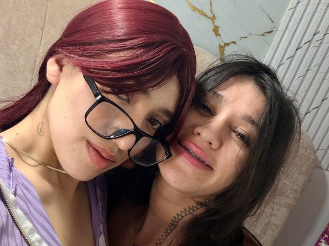 AbbyAndMichelross with red hair and glasses, leaning on AbbyAndMichelross with black hair, both smiling, showing off their tits and pussy tattoos, on a beige couch.