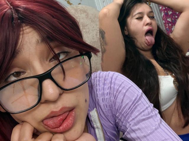 AbbyAndMichelross with red hair, glasses, and pouty lips in foreground, tongue out, arms up, brown hair, white bra, both showing playful expressions. AbbyAndMichelross's tits visible.