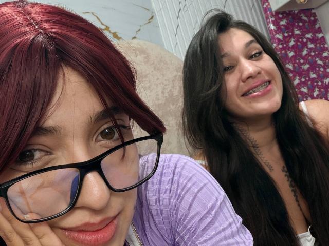 AbbyAndMichelross with red hair, glasses, and pink shirt, and AbbyAndMichelross with black hair, smiling, showing tits in a photo. AbbyAndMichelross has tattoos on her neck.