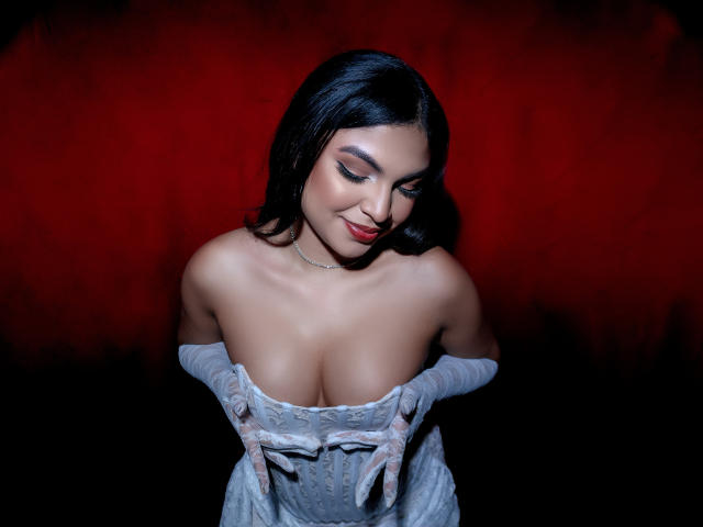 Melody Alvarez mit dunklem Haar, rotem Lippenstift und weißen Handschuhen, zieht ein Spitzenkleid herunter, um ihre großen, nackten Brüste vor einem roten Hintergrund zu zeigen..