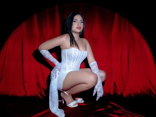 Melody Alvarez mit schwarzen Haaren, einem weißen Spitzen-Korsett und Handschuhen, hockt, zeigt ihr Gesäß, trägt weiße High Heels und steht vor einem roten Vorhang, sieht verführerisch aus..