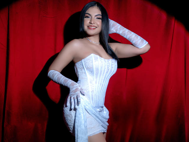 Melody Alvarez, in einem weißen, trägerlosen, korsettartigen Kleid mit Spitzenhandschuhen, lächelnd, eine Hand am Hüfte, vor einem roten Hintergrund, wobei ihre Brüste, ihr Hintern und ihr hübsches Gesicht zu sehen sind..