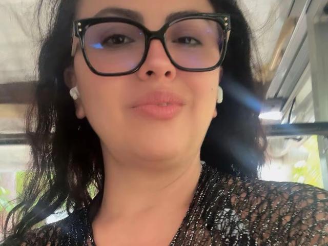 CharlotteDevereaux with black glasses, wearing a sheer black top, has puffy pink lips, dark wavy hair, and white earrings, showing a subtle smile, her nose is centered.