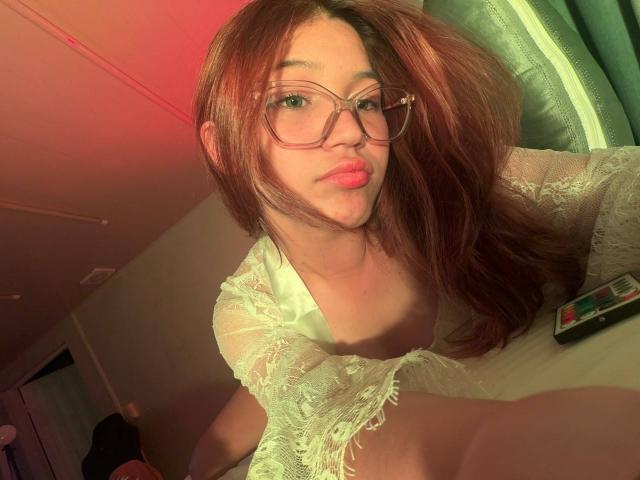 ElodieGardner with red hair, glasses, and a white lace top, showing her small tits, sitting on a bed, looking at the camera, with a phone on the right.