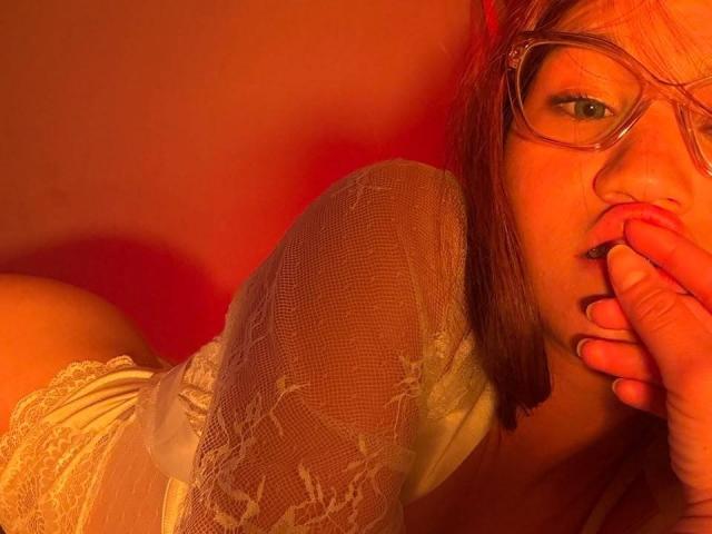 ElodieGardner with glasses, biting her finger, wearing white lace, has red hair, and is looking at the camera with a seductive expression, in warm red lighting.