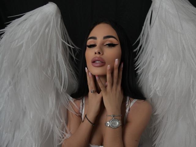 CharlotteAria with white angel wings, long black hair, thick eyebrows, heavy eyeliner, pink lipstick, hands on her face, pink nails, silver watch, black background, and white feather wings.