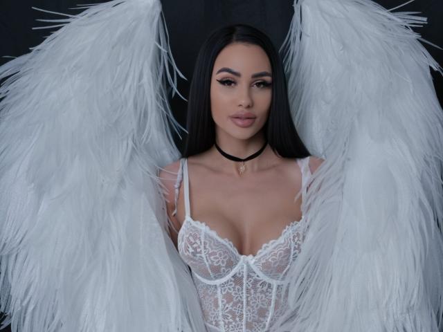 CharlotteAria with white angel wings, wearing a lace white corset, black choker, and long black hair, has large tits, full lips, and a cute face, looking directly at camera.