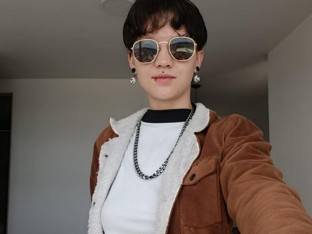 LissyLuxe, con cabello corto castaño, usando gafas de sol tipo aviador reflectantes, una chaqueta de ante marrón con forro de polar blanco, una camiseta blanca, un collar negro y aretes negros, mostrando una sonrisa segura..