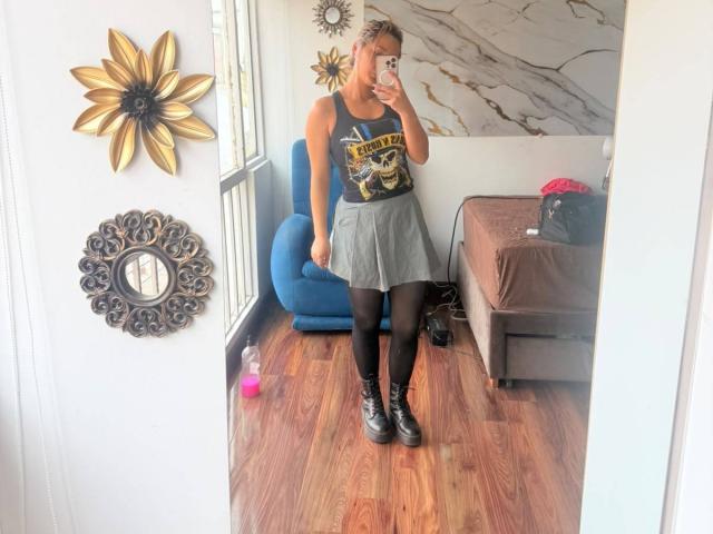 ElenaSaenz has short blonde hair, wears a black tank top, gray skirt, black tights, and black boots, standing in front of a mirror, holding a phone.