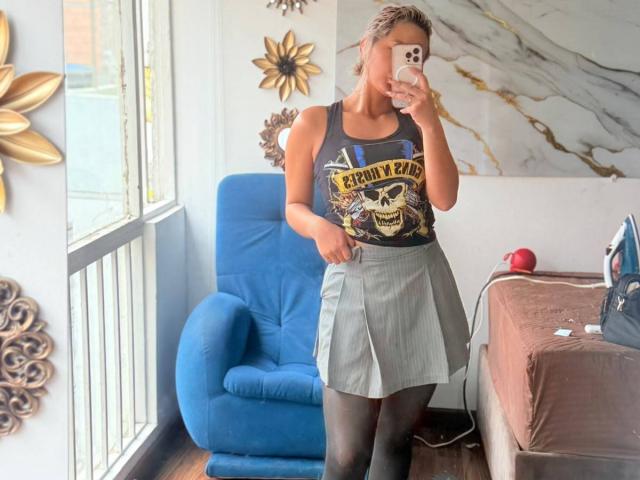 ElenaSaenz with small tits, wearing a black "Electric Priest" tank top, gray skirt, black tights, taking a mirror selfie, standing in a room with blue chair.