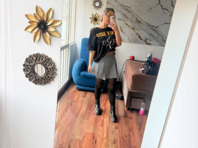 ElenaSaenz with short gray hair, wearing a black "Guns N' Roses" shirt, gray skirt, black tights, and black boots, taking a mirror selfie in a wooden room.