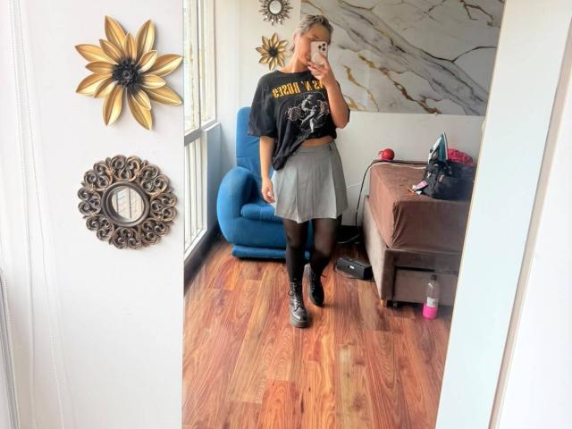 ElenaSaenz in a black shirt, gray skirt, black tights, and black boots, standing in front of a mirror, holding a phone, showing her ass, in a wooden room.
