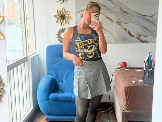 ElenaSaenz with short gray hair, wearing a skull tank top, gray skirt, black tights, and holding a phone, standing in a room with a blue chair and brown bed.