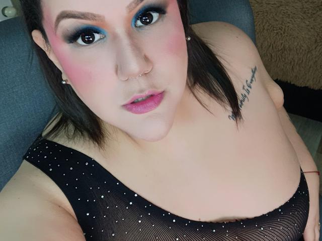 GiiannaMorrys with blue eyeshadow, pink blush, and pink lipstick, wearing a black mesh top, showing her tits, nose ring, and tattoo on her right shoulder.