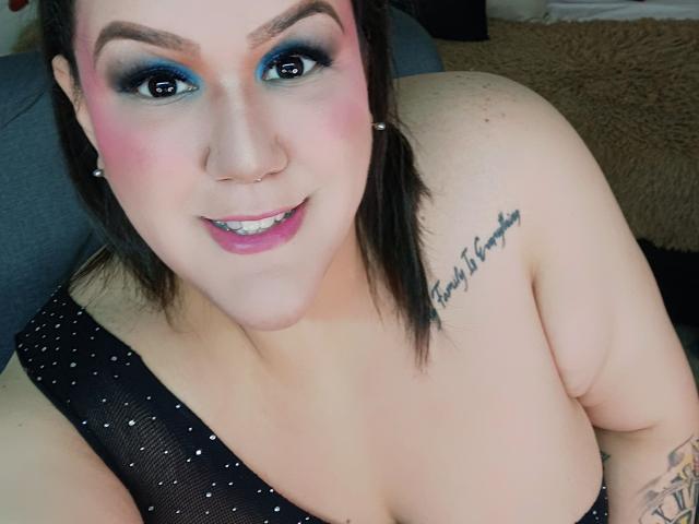 GiiannaMorrys with blue eyeshadow, pink blush, nose ring, black mesh top, showing her tits, tattoo on right shoulder, smiling, black hair, and a small pink lipstick.