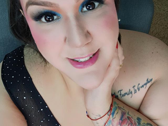 GiiannaMorrys with blue eyeshadow, pink blush, and pink lipstick, smiling, hand on cheek, black polka-dot top, tattoo on shoulder, red bracelet, and colorful arm tattoo.
