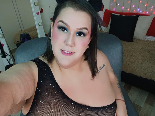 GiiannaMorrys with pink eyeshadow, blue eyeshadow, black mesh top, small nose ring, tattoo on right arm, taking selfie, smiling, pink lipstick, visible arm stretch, brown hair.