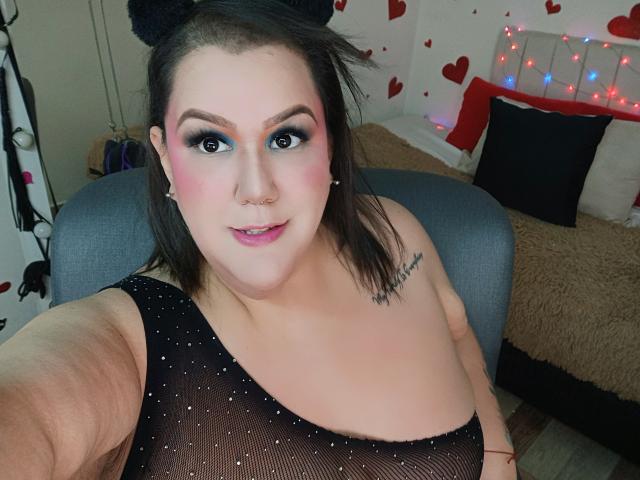 GiiannaMorrys with pink eyeshadow, blue eyeliner, and black mesh top showing her tits, a nose ring, and tattoo, taking a selfie in a bedroom with heart decorations.
