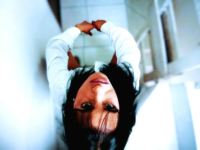 NinnaDesire's face upside down, dark hair, green eyes, white shirt, arms crossed, looking at camera, bright lighting, soft focus, slightly blurred background, tilted perspective.