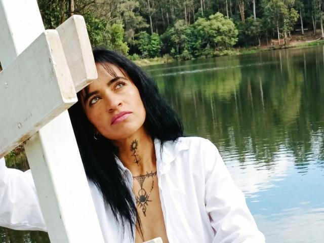 NinnaDesire with black hair, white shirt, tattoos on neck, leaning on white fence, water and forest in background, looking up, pink lips, hoop earrings, serious expression.